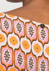 Multikleuren stof met bloem- en geometrische patronen in oranje, roze en bruin op een lichte achtergrond met een klein logo-detail.