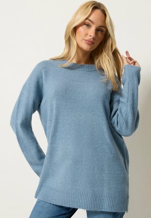 Kvinde med langt blondt hår iført en løs lyseblå sweater og blå bukser, poserer med en hånd, der rører ved håret mod en ensfarvet baggrund.