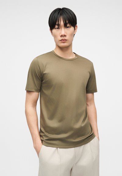 Νεαρός άνδρας με μαύρα μαλλιά, φοράει ένα μπλουζάκι σε χρώμα ελιάς με στρογγυλή λαιμόκοψη και ανοιχτό μπεζ παντελόνι, στέκεται με τα χέρια στις τσέπες μπροστά σε μια μονόχρωμη επιφάνεια.