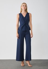 Haut sans manches bleu marine avec un décolleté en V et un design torsadé à la taille, associé à un pantalon large bleu marine. Texture de tissu lisse et matte.