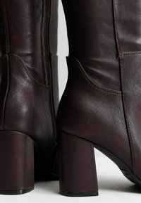 Botines de cuero burdeos oscuro con tacón grueso, con cremallera lateral, textura suave y detalles curvos a lo largo de las costuras.