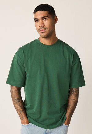 Mand med kort hår og tatoveringer på armene iført en ensfarvet grøn oversized T-shirt og lyseblå jeans, hænderne i lommerne, neutral baggrund.