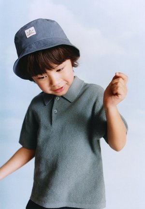 Malen otrok z modrim klobukom v vedru in teksturirano sivo polo majico, ki gleda navzdol z dvignjenimi rokami na ravni svetli podlagi.