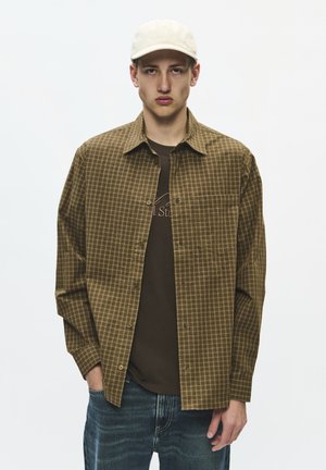 Jeune homme portant une casquette beige, une chemise marron à carreaux boutonnée sur un t-shirt foncé et un jean bleu, debout devant un fond uni.
