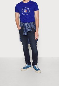 Camiseta de algodón azul con logo circular blanco, camisa de cuadros atada a la cintura, vaqueros oscuros y zapatillas azul con estampado.