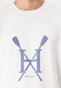 Hvit hettegenser med en blå grafikklogo med kryssede årer og merkevarenavnet "Hackett" under. Klassisk rund halslinning og mykt stoff.