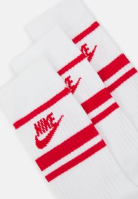 Bijele atletske čarape s crvenim prugama i crvenim Nike logotipom. Rebrasta tekstura, prikazane tri pare. Uzorak pruga oko vrha.