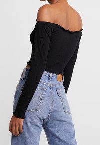 Černý top s odhalenými rameny a dlouhými rukávy, texturovaná tkanina a volánkové okraje, v kombinaci s vysokým pasem světle modrými džíny se zadními kapsami.