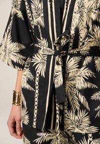Tessuto con motivo di palme in nero e beige, con maniche ampie, cintura in vita e perline decorative. Materiale in cotone con texture liscia.