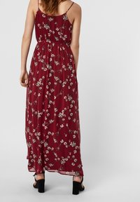 Femme debout portant une robe maxi sans manches bordeaux à imprimé floral blanc, associée à des sandales à talons noirs, vue de dos.