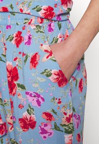 Pantalon floral bleu avec une taille élastique. Présente des fleurs rouges, roses et vertes. Inclut un détail de poche latérale avec une texture douce.