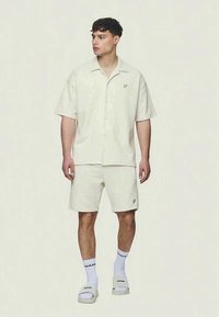 Camicetta chiara beige a maniche corte con bottoni e pantaloni corti coordinati, entrambi con una texture. Indossata con calzini bianchi e sandali; logo sulla tasca della camicia.