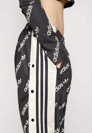 Czarne dresowe spodnie Adidas z białymi paskami i wzorem logo, noszone z dopasowaną bluzką z długim rękawem, pokazującą rękę w kieszeni.