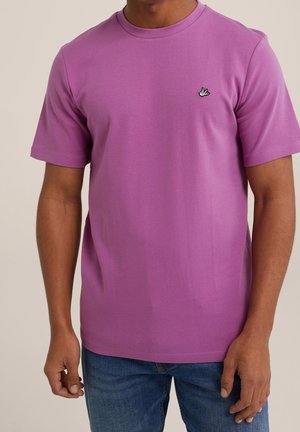 Man draagt een effen paarse korte mouwen T-shirt met een klein wit banaanlogo op de borst en een blauwe spijkerbroek, staand tegen een neutrale achtergrond.