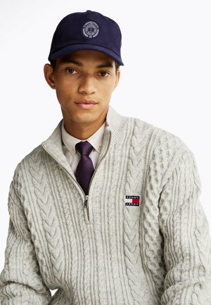 Navy baseball cap with logo, grey cable-knit sweater with high collar, and white dress shirt underneath. Notable zip detail and textured pattern.