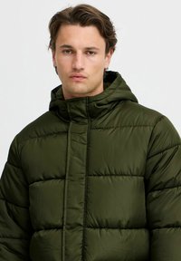 Puffet, mørkegrønn hettegenser med quiltet design, med vertikale sømmer og hel glidelås foran. Myk, jevn tekstur.