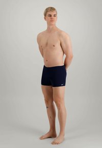 Tengerészkék úszónadrág, rugalmas derékkal és fehér Nike logóval, álló férfi modellen viselve. Simább anyag, minimalista dizájn, combközépig érő hossz.