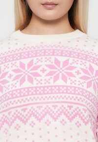 Woman wearing white knit sweater with pink geometric and floral patterns, shown from chin to chest.