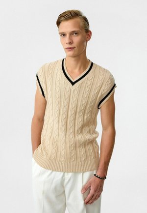 Beige gebreide mouwloze trui met een V-hals, voorzien van een kabelpatroon en zwarte bies langs de halslijn en armsgaten.