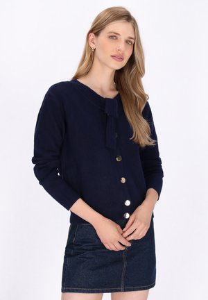 Navy button-up cardigan with a collar tie. Features gold-tone buttons, ribbed cuffs, and a soft texture. Paired with a denim skirt.