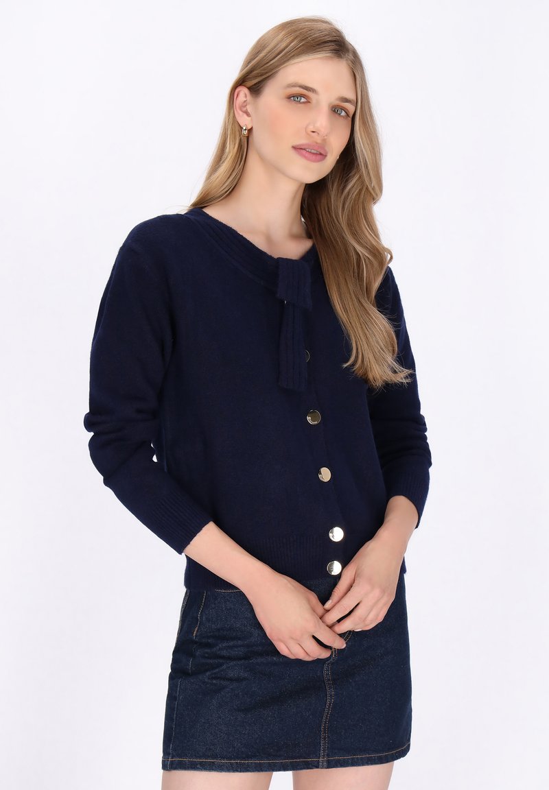 Marineblauwe cardigan met knopen en een kraagstrik. Beschikt over goudkleurige knopen, geribbelde manchetten en een zachte textuur. Gecombineerd met een denim rok.