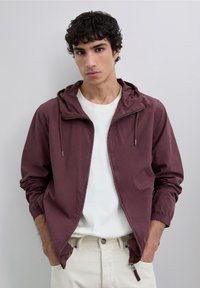 Ung mand med mørkt krøllet hår iført en hvid t-shirt og en åbnet maroon hættejakke, hænderne i lommerne, neutral baggrund.