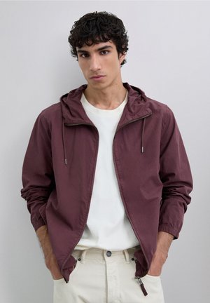 Joven con cabello rizado oscuro que lleva una camiseta blanca y una chaqueta con capucha burdeos sin cerrar, con las manos en los bolsillos, fondo neutro.