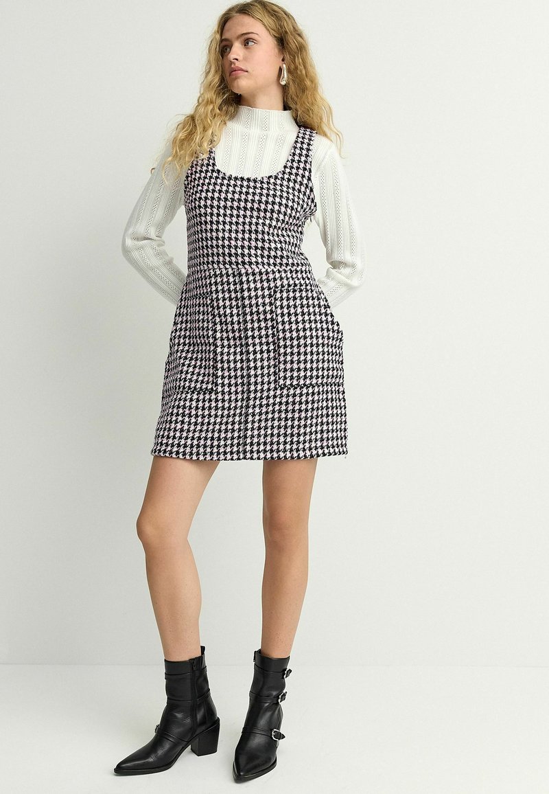 Melnā un baltā houndstooth rakstā raiba, bez piedurknēm, pieguļošā vidukļa kleita ar sānu kabatām, apvienota ar melniem potīšu zābakiem.