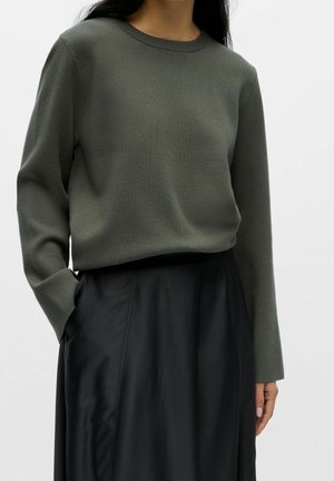 Žena v nošeném khaki zeleném svetru s dlouhými rukávy zasunutém do černé sukně s vysokým pasem, stojící s jednou rukou v kapse.