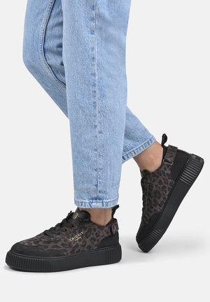 Voeten met zwarte en bruine luipaardprint sneakers met zwarte dikke zolen en lichtblauwe spijkerbroek tegen een effen achtergrond.