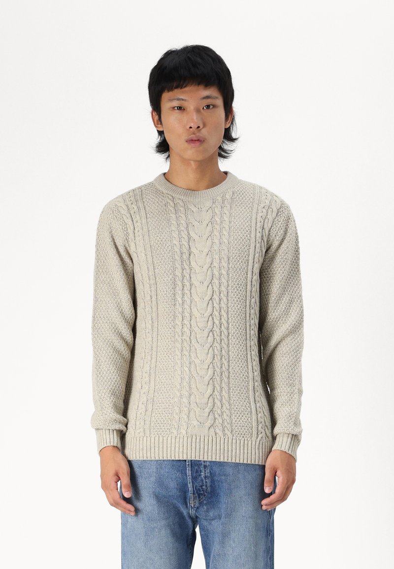 Suéter de punto beige con patrones texturizados y cuello redondo, combinado con jeans de mezclilla azul. Tejido suave y cálido con puños acanalados.