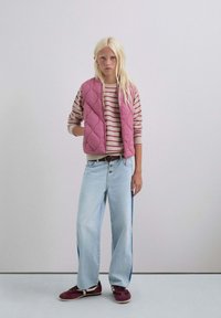 Chaleco acolchado rosa sobre un suéter a rayas en crema y rojo, combinado con jeans de pierna ancha en azul claro y zapatillas burdeos con acentos en blanco.