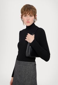 Melnā ribiņu turtleneck džemperis savienots ar pelēku hounds-tooth svārku. Modelis tur elegantu melnu somu ar strukturētu dizainu.