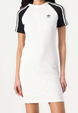 Femme portant une robe Adidas blanche avec des manches et des bandes d'épaules noires, affichant le logo Adidas sur la poitrine.