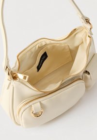 Petit sac à main couleur crème avec fermetures éclair et anneaux dorés, ouvert pour montrer une poche intérieure unique et une doublure en tissu avec étiquette de la marque.