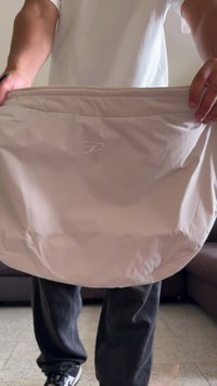 Saco de tecido bege claro com uma superfície texturizada, apresentando um logotipo subtil e uma forma arredondada, segurado por uma pessoa vestindo roupas casuais.