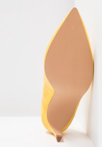 Escarpin en daim jaune avec un talon aiguille, un bout pointu, une tige lisse et une semelle texturée marron clair. Dispose d'un talon fin pour une silhouette élégante.