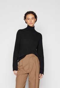 Schwarzer Rollkragenpullover mit langen Ärmeln, aus weichem Material; kombiniert mit hoch taillierten, plissierten hellbraunen Hosen.