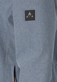 Light blue jacket with a textured fabric, featuring a black logo patch on the sleeve and a zippered pocket. Smooth seam detailing.