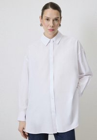 Biała, oversize koszula zapinana na guziki z klasycznym kołnierzykiem, długimi rękawami i gładką teksturą materiału. Stylizowana z ciemnymi dżinsami.
