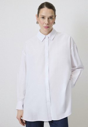Biała, oversize koszula zapinana na guziki z klasycznym kołnierzykiem, długimi rękawami i gładką teksturą materiału. Stylizowana z ciemnymi dżinsami.