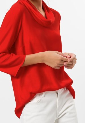 Vrouw draagt een losse rode top met lange mouwen en een brede kraag en witte broek, handen in elkaar geslagen ter hoogte van de taille tegen een effen achtergrond.