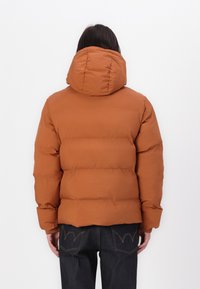 Brun polstret jakke med hætte, med vandret syninger og elastiske manchetter, parret med mørk denim jeans. Glat tekstur.