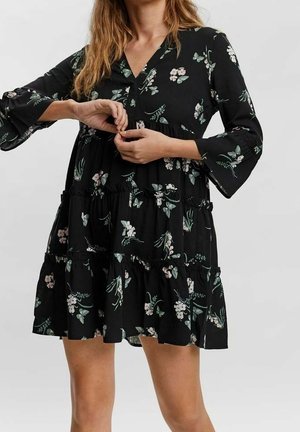 Femme portant une robe noire à fleurs avec un col en V, des manches longues et une jupe à volants, boutonnant la taille à l'avant.
