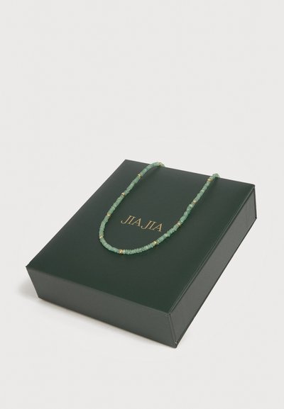 Πράσινο κολιέ με χάντρες και χρυσά διαχωριστικά που ακουμπά σε ένα σκούρο πράσινο ορθογώνιο κουτί με χρυσή επιγραφή "JIA JIA".