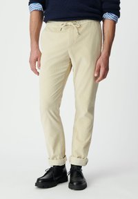 Pantalones de pana color crema con cintura ajustable, corte entallado, puños enrollados y combinados con zapatos negros con cordones. Detalle de tela texturizada.