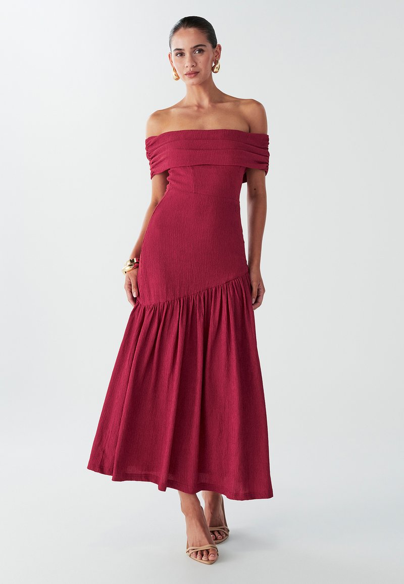 Robe maxi bordeaux à épaules dénudées avec tissu texturé, corsage ajusté et jupe froncée. Accessoires : grandes boucles d'oreilles créoles.