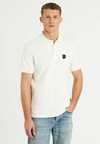 Valge polo särk, mis on valmistatud tekstureeritud kangast, ribikrae ja lühikeste varrukatega. Vasakul rinnal on väike must logo.