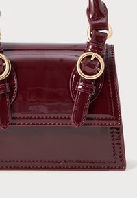 Sac à main en cuir verni bordeaux avec quincaillerie dorée, de forme rectangulaire, doté de deux poignées et de coutures visibles.