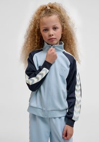 Noor tüdruk lokkis juustega, kandes helesinist ja tumesinist dressipluusi, hoides käes jope tõmblukku ühevärvilisel taustal.